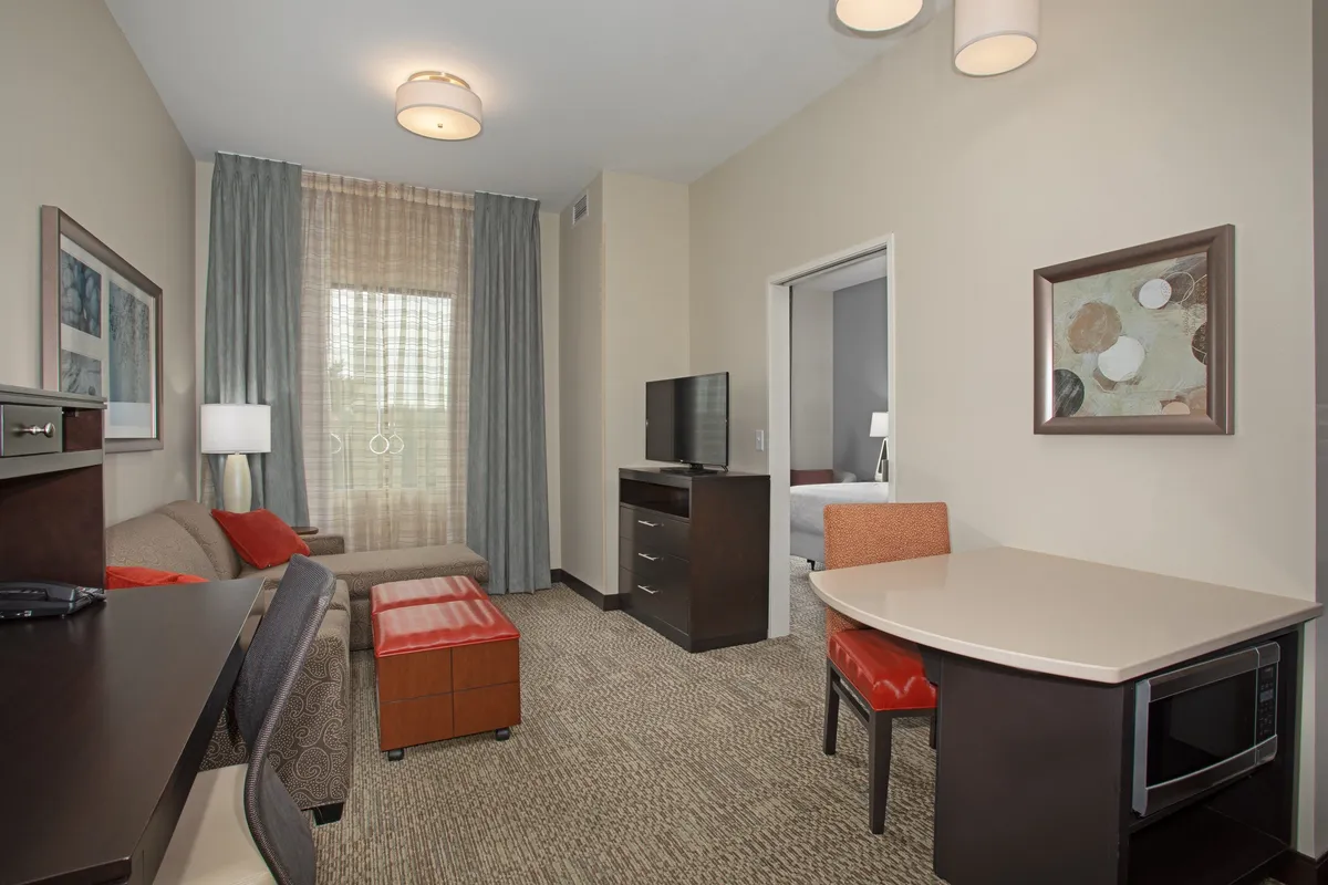 The image shows a stylish, comfortable living area in a one-bedroom condo, featuring a plush sofa facing a flat-screen television, a cozy dining table for two, bright natural light streaming through a large window, and a door leading to the private bedroom. The space is inviting, perfect for relaxing, dining, or working, and exudes a calm, homey atmosphere ideal for a couple or solo traveler.
