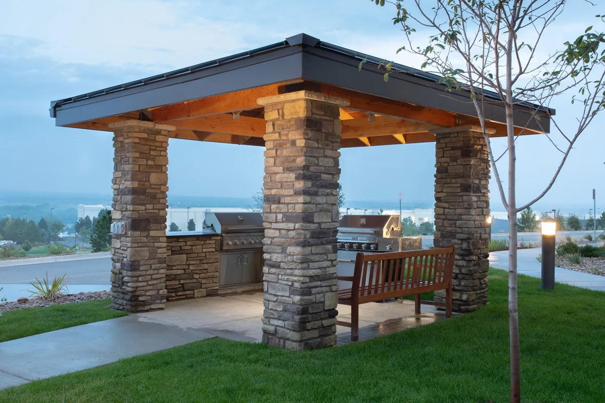 This exterior image showcases sturdy stone pillars framing a welcoming outdoor barbecue area with grill stations and a wooden bench. The expansive patio is set against a backdrop perfect for gatherings, socializing, and enjoying the fresh air. The casual ambiance is accentuated by soft evening lighting, evoking the spirit of sunset barbecues and starlit community events.