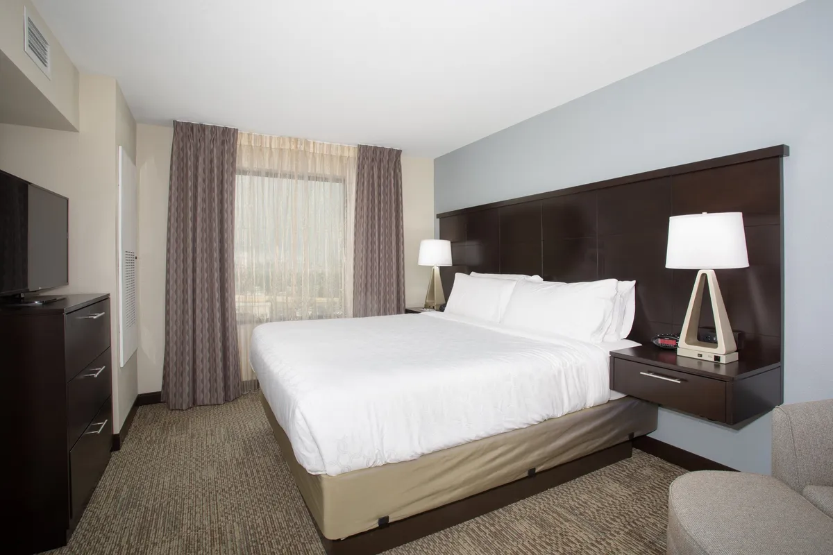 A cozy bedroom scene featuring a Queen bed neatly made with plush bedding, flanked by a nightstand and a window letting in soft natural light. A wall-mounted television faces the bed, creating an inviting space for relaxation, reading, or watching TV. The overall mood is peaceful and serene, with subtle decor touches.