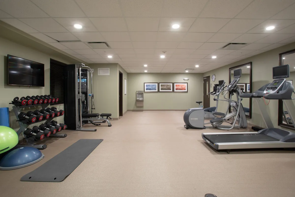 A motivating fitness center filled with a variety of modern exercise equipment, including a dumbbell rack, treadmills, elliptical machines, and exercise mats. Wall art adds an energetic touch, and the space is designed for both individual and group workouts.