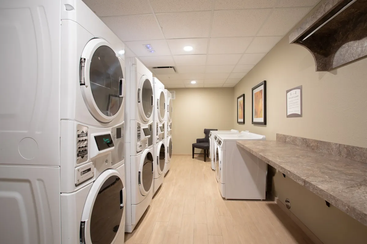 A clean, well-lit laundry room features multiple modern laundry machines, a spacious folding counter, and a comfortable seating area, providing guests with a practical space for taking care of laundry needs during longer stays.